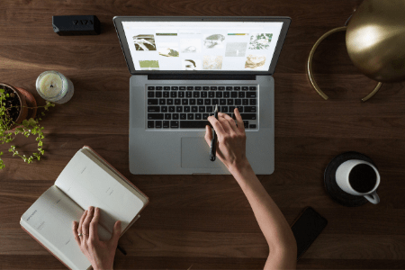 Photo of a desk from above with a laptop, pen, notebook and coffee mug