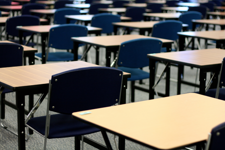 Photo showing an empty exam hall full of rows of small wooden desks and blue chairs